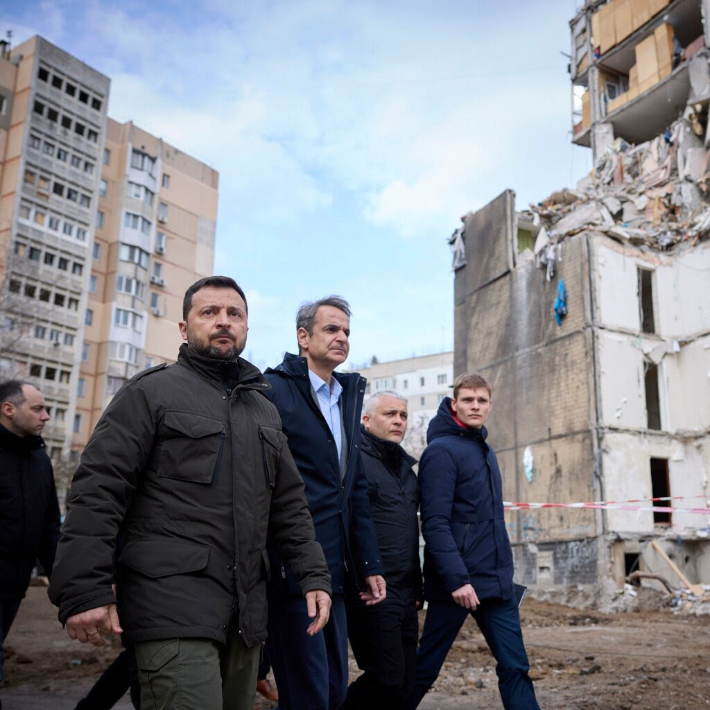Ukrainian President Volodymyr Zelensky and Greek Prime Minister Kyriakos Mitsotakis walk in front of damaged buildings.