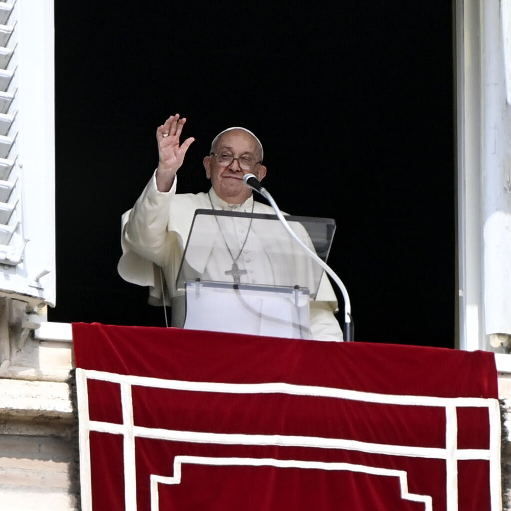 Pope Francis, in white, gestures from an open window, with a lectern and microphone in front of him.