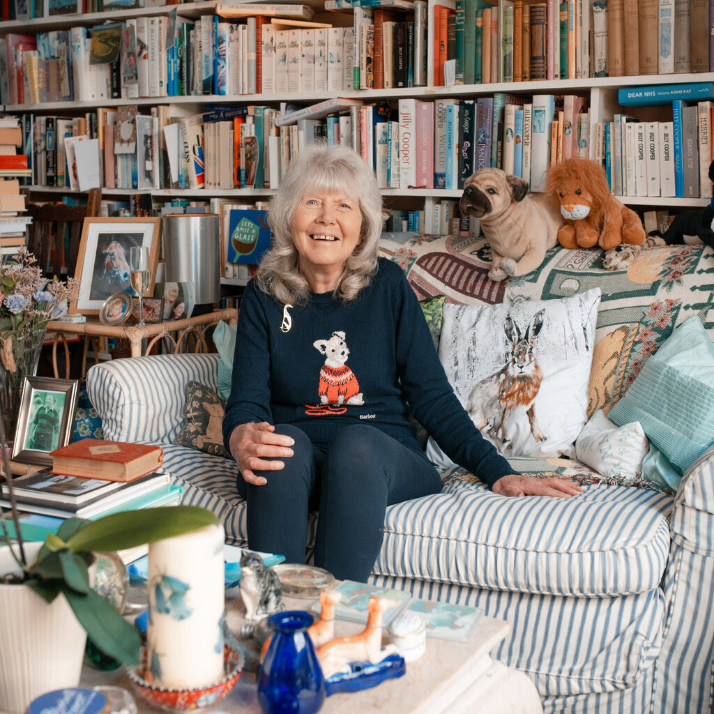 A woman with gray hair sits on a stripy sofa, laughing, with shelves of books behind her.