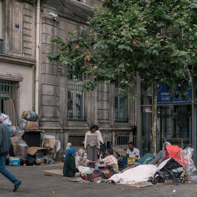 A group of people, their possessions around them, sit on the sidewalk next to a building.   