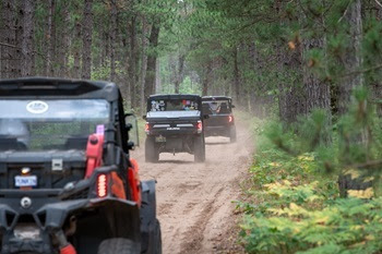 three black off-road vehicles ride single-file down a tree-lined, dirt trail in the Manistee National Forest