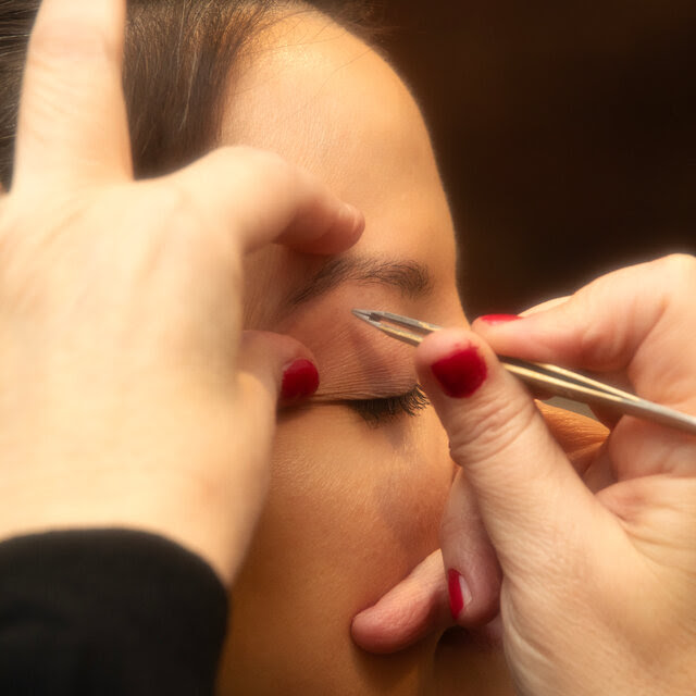 A woman has her eyebrows tweezed by another woman with red painted nails.