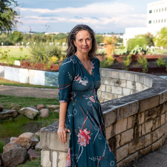 Natalie Vallot, wearing a green floral dress, leaning against a low brick wall with a hospital in the background. 