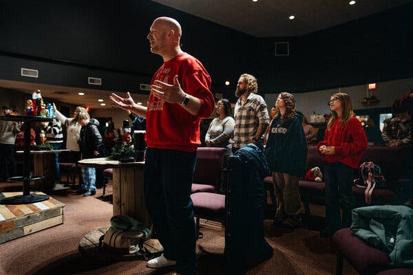 Billy Holden, vistiendo una sudadera roja, levanta las manos mientras canta en un servicio religioso.