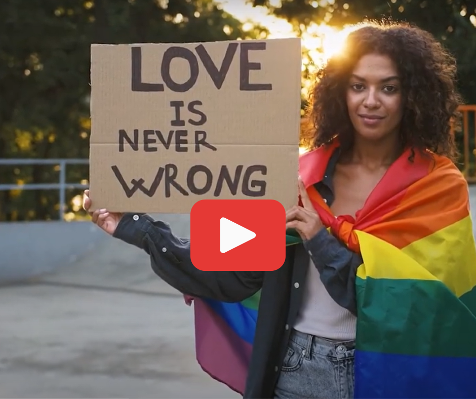 Person with a pride flag wrapped around their shoulders holding a sign that reads: Love is never wrong.