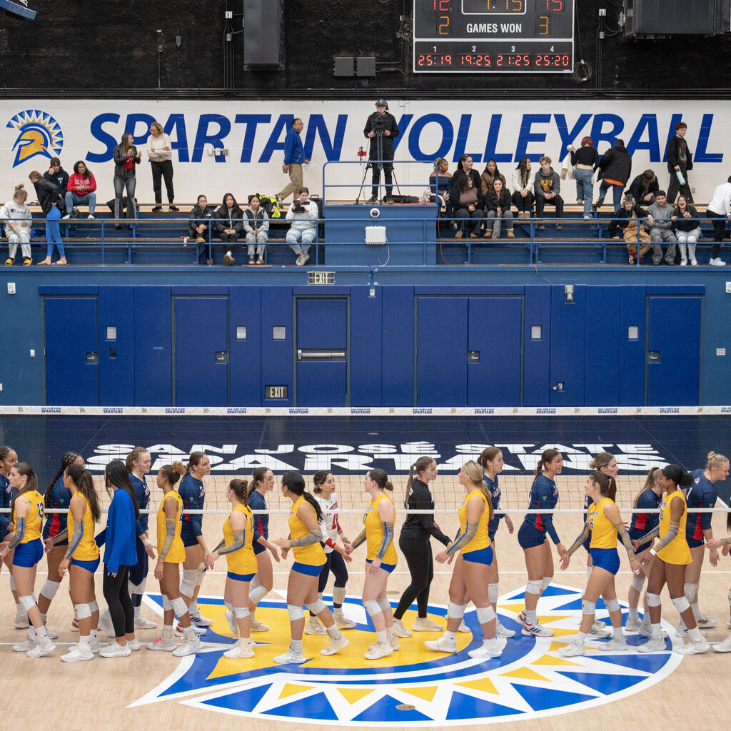 Female volleyball players line up on either side of a volleyball net in a gym. A sign on the back wall says “Spartan Volleyball.”