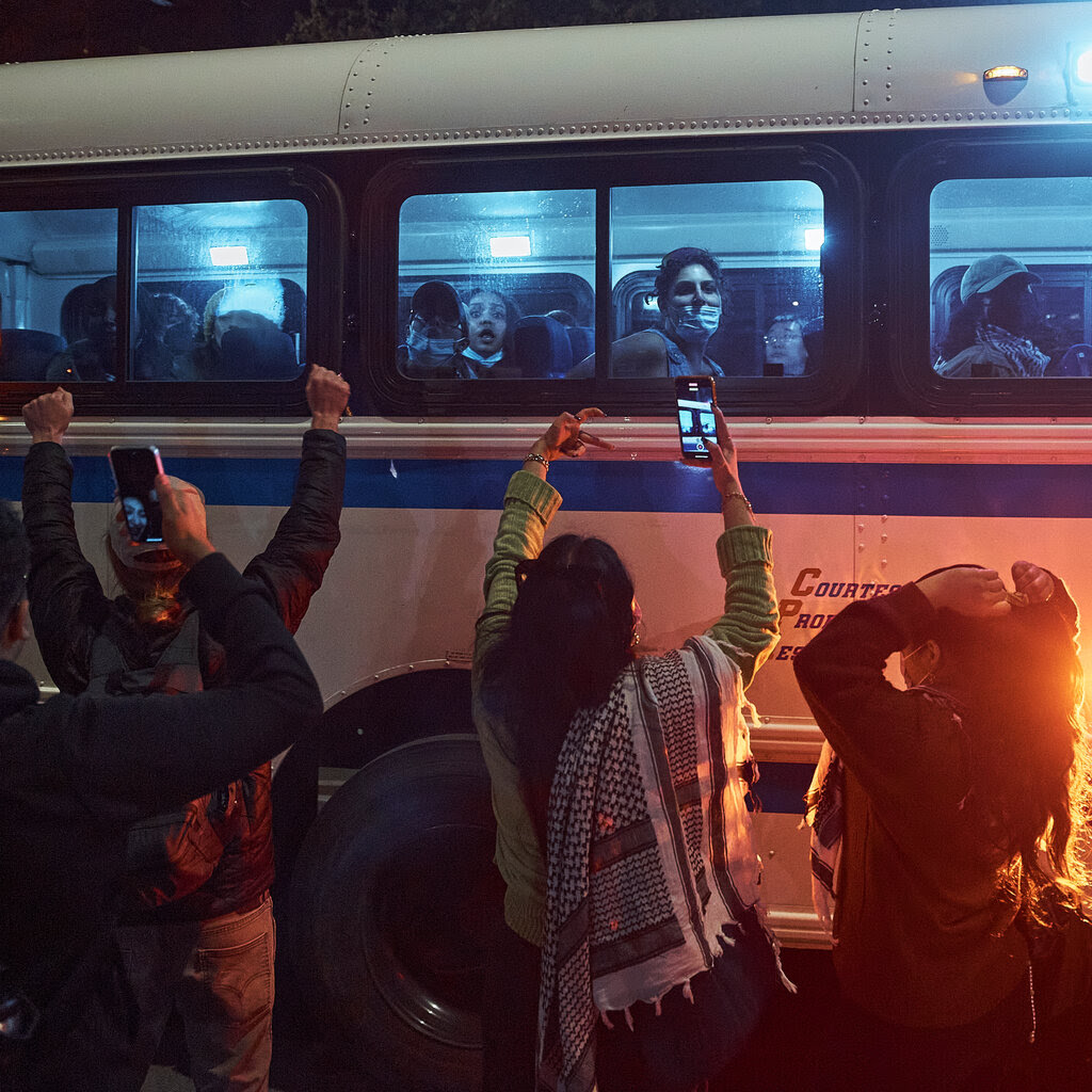 Protesters stare out the windows of a police bus as other people outside cheer for them.