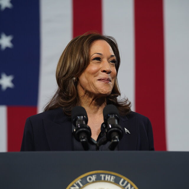 Kamala Harris stands smiling behind a lectern, with a large American flag behind her.