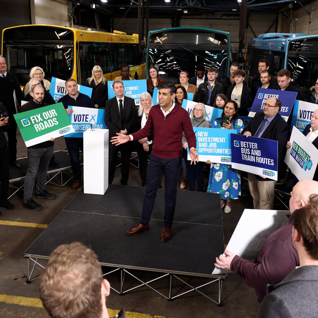 Rishi Sunak stands on a raised platform in a bus depot surrounded by a crowd holding signs supporting the Conservative Party.