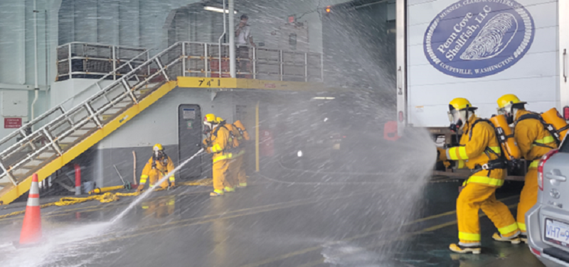 Several people in firefighting gear spraying water on out of hoses on the car deck of a ferry