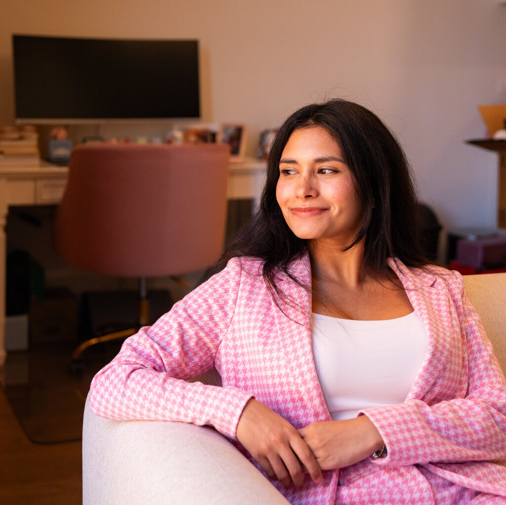 A woman in a pink houndstooth blazer smiles as she sits in a chair.