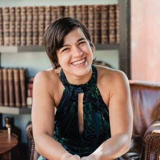 The photo portrays a woman with short brown hair, seated, wearing a green-and-black flocked velvet top, with hands folded over her knees. She is wearing green nail polish.