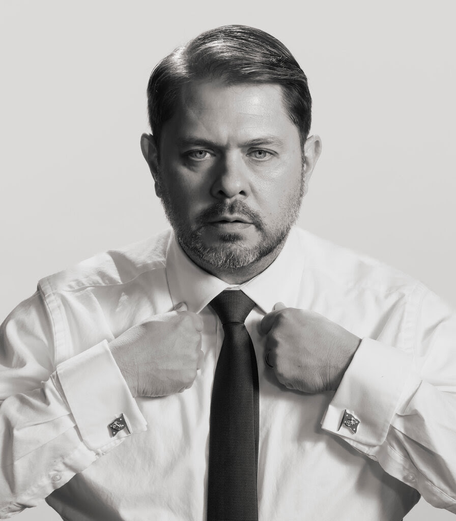 A black-and-white portrait of Senator Ruben Gallego in a white shirt and tie. 