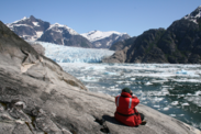  Harbor seal researcher in orange jumpsuit sits with binoculars on a rockface bank of ice capped water