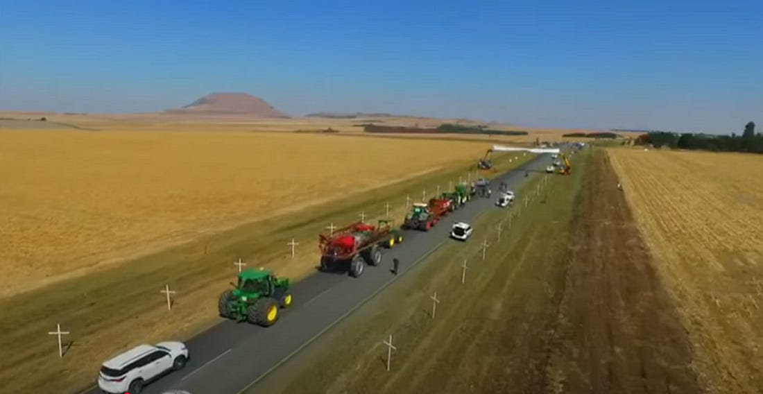 File di croci bianche si vedono su entrambi i lati di una strada rurale in Sudafrica. Trattori e automobili percorrono il centro della carreggiata, con campi su entrambi i lati.