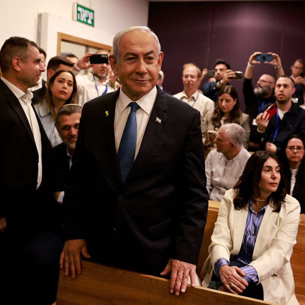 Benjamin Netanyahu, prime minister of Israel, stands smiling and wearing a suit in a crowded room, with his hands touching the wooden backrest of a bench. 