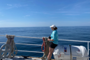 Abby Lucas in dark shorts, a light blue long-sleeve shirt, and a baseball hat holding a camera at the railing of a ship on a clear day with calm water