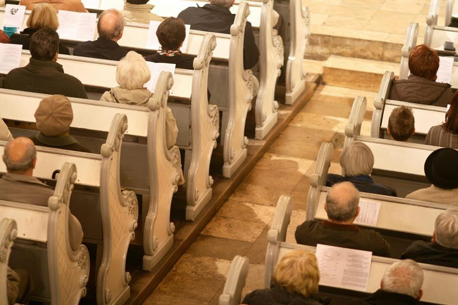 Several rows of white church pews, with about a dozen people sitting on them.