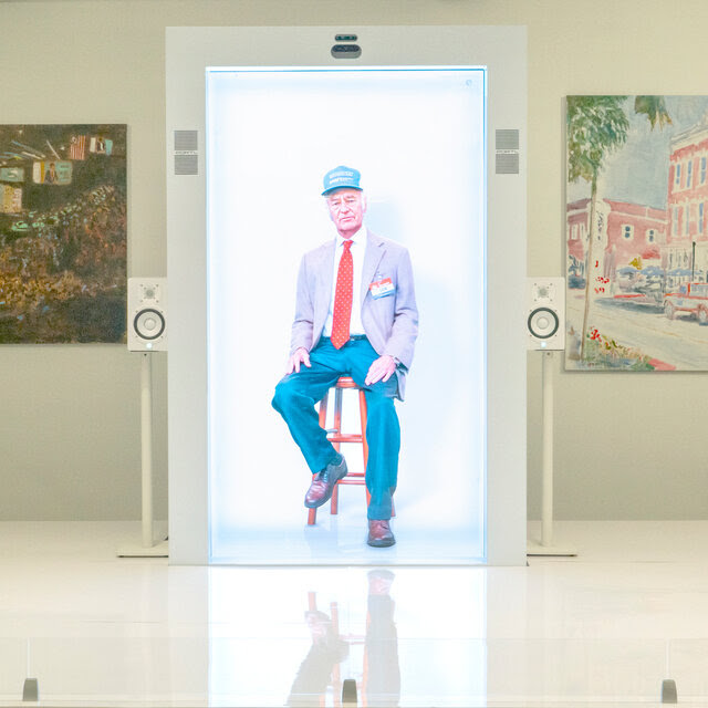 A hologram of Sam Walton, sitting on a stool and wearing a blue cap, light colored blazer and blue pants, on a platform with paintings on either side.