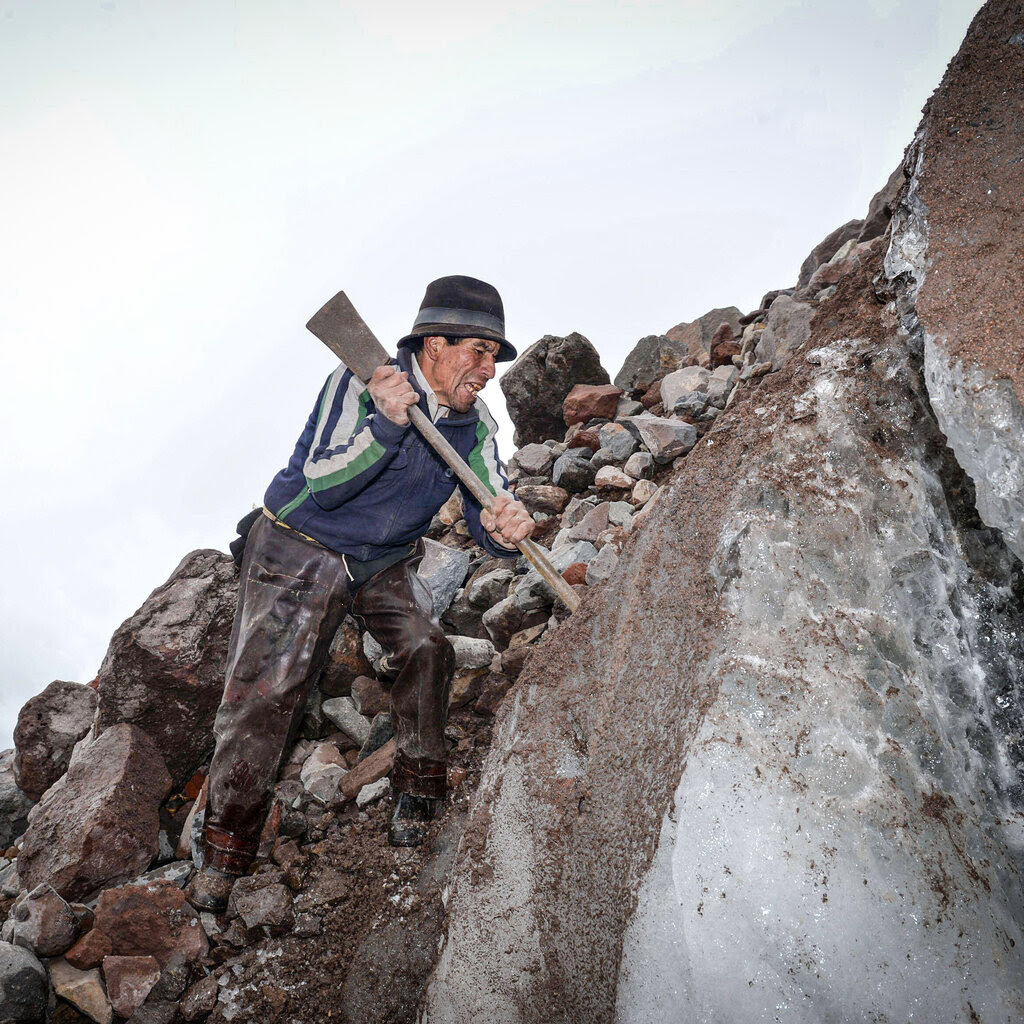 Baltazar Ushca wearing a sweater, work pants and a hat, hoists a pickax over his shoulder while balancing hi on the side of an icy mountain.