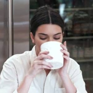 Person drinking from a white cup in a kitchen, wearing a casual white shirt