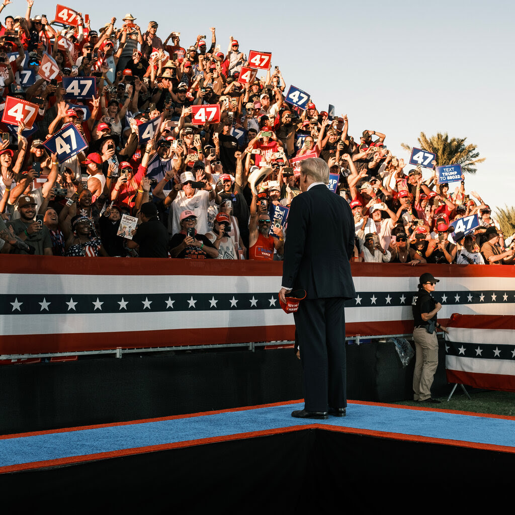 Donald Trump stands on a walkway to a stage, staring at an outdoor crowd of cheering supporters.