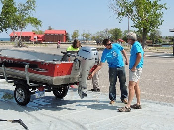 At a landing blitz, two men standing near the motor of a trailered small boat in a parking lot. A woman and a child are standing behind the boat.