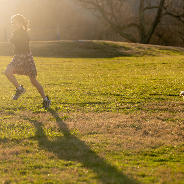 A young girl runs across a grassy lawn, trailed by a small dachshund.