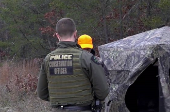 a male conservation officer in brown camo uniform with POLICE on the back talks to a hunter wearing orange cap outside a camo ground blind