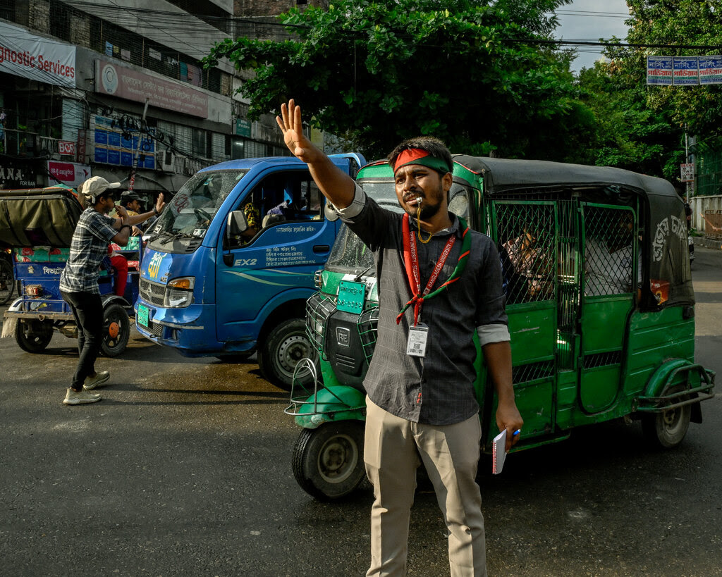 A person wearing a red and green headband stands in the street with a whistle in their mouth and raises their hand. 