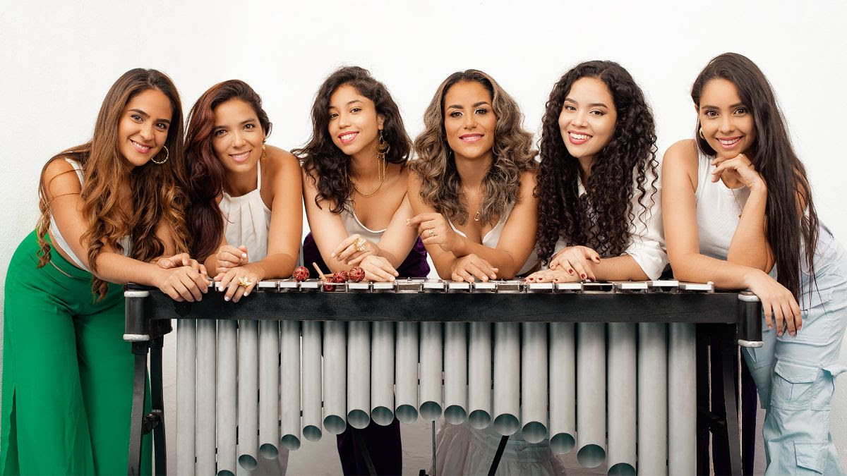 Six women with long hair lean smiling on a vibraphone