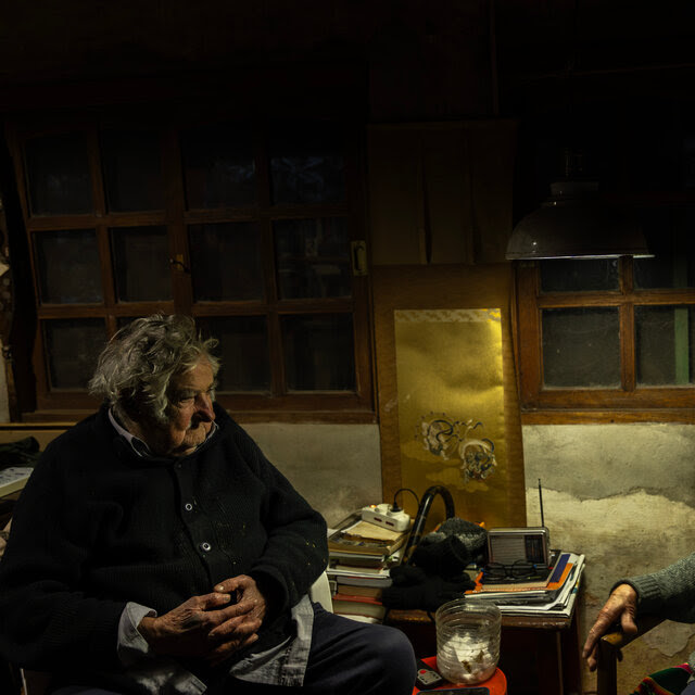 José Mujica and Lucía Topolansky sitting in chairs in their home in dim lighting. She smiles at him.