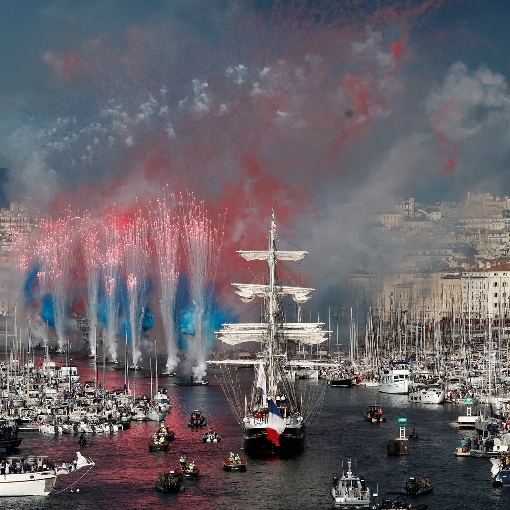 A three-masted sailing ship arriving at the port of Marseille as flares erupt. 