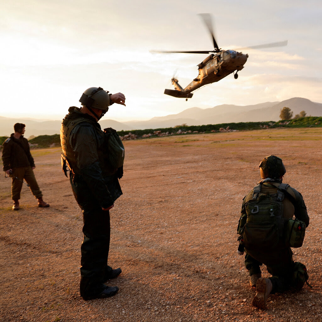 A Blackhawk helicopter hovers above a patch of dirt as four soldiers look on.