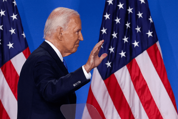 Joe Biden, a balding white man wearing a dark blue suit, waves hand near American flags.