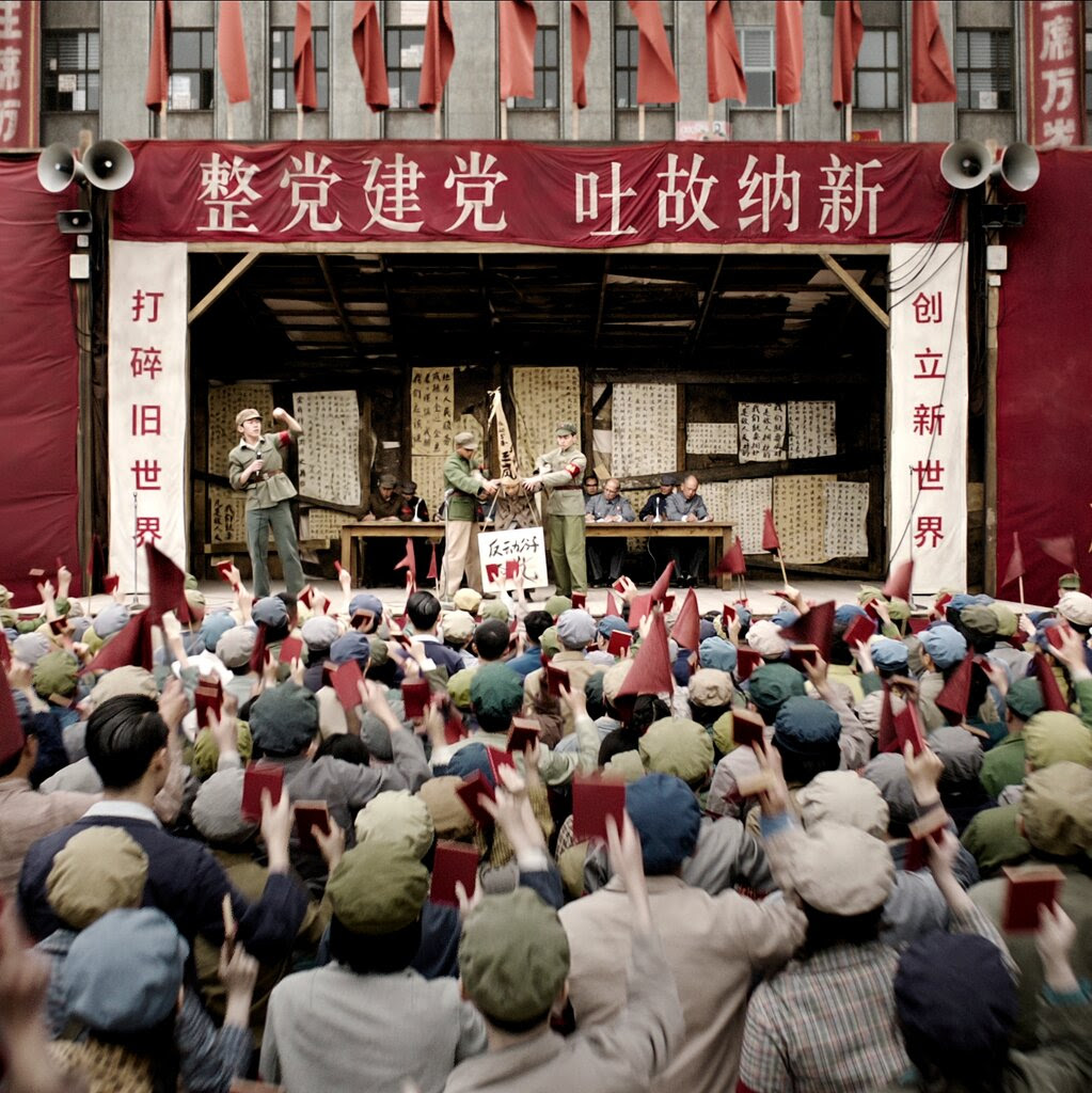 A crowd faces a stage, some waving red flags. Men in uniforms are on the stage, including a man holding a microphone.
