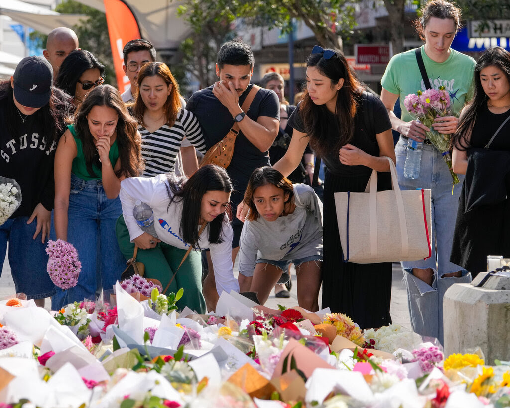 People gather at a street memorial, some adding bouquets to a huge floral display on the ground.