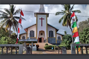 Trees surround a church building behind an open gate. There are colourful flags on either side of the gate.