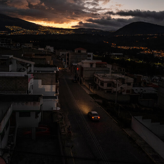 A car rides along a darkened street with a view of mountains and lights in the background.