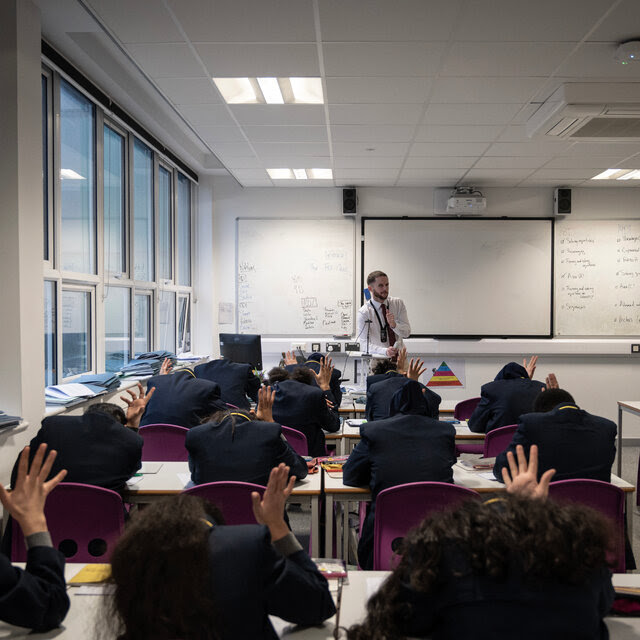 A class of students sitting at desks with their heads bowed and one hand raised, while a teacher stands at a whiteboard at the front.