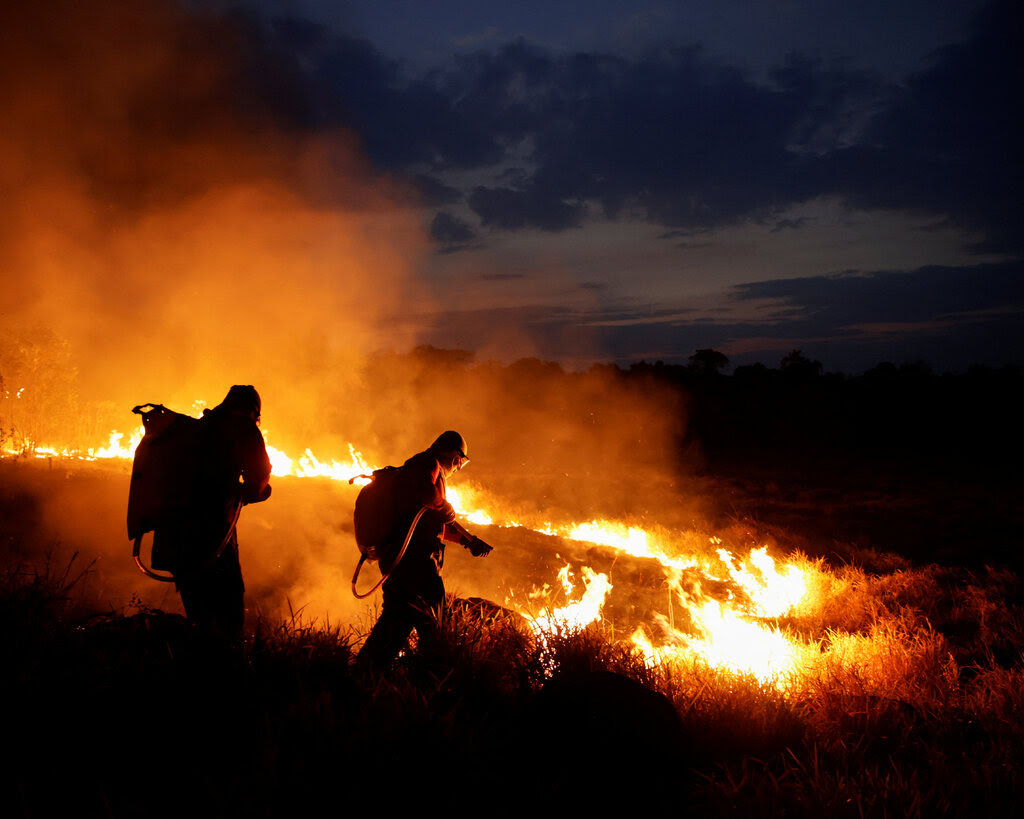 Two firefighters battle a blaze amid brush on a hillside at night.