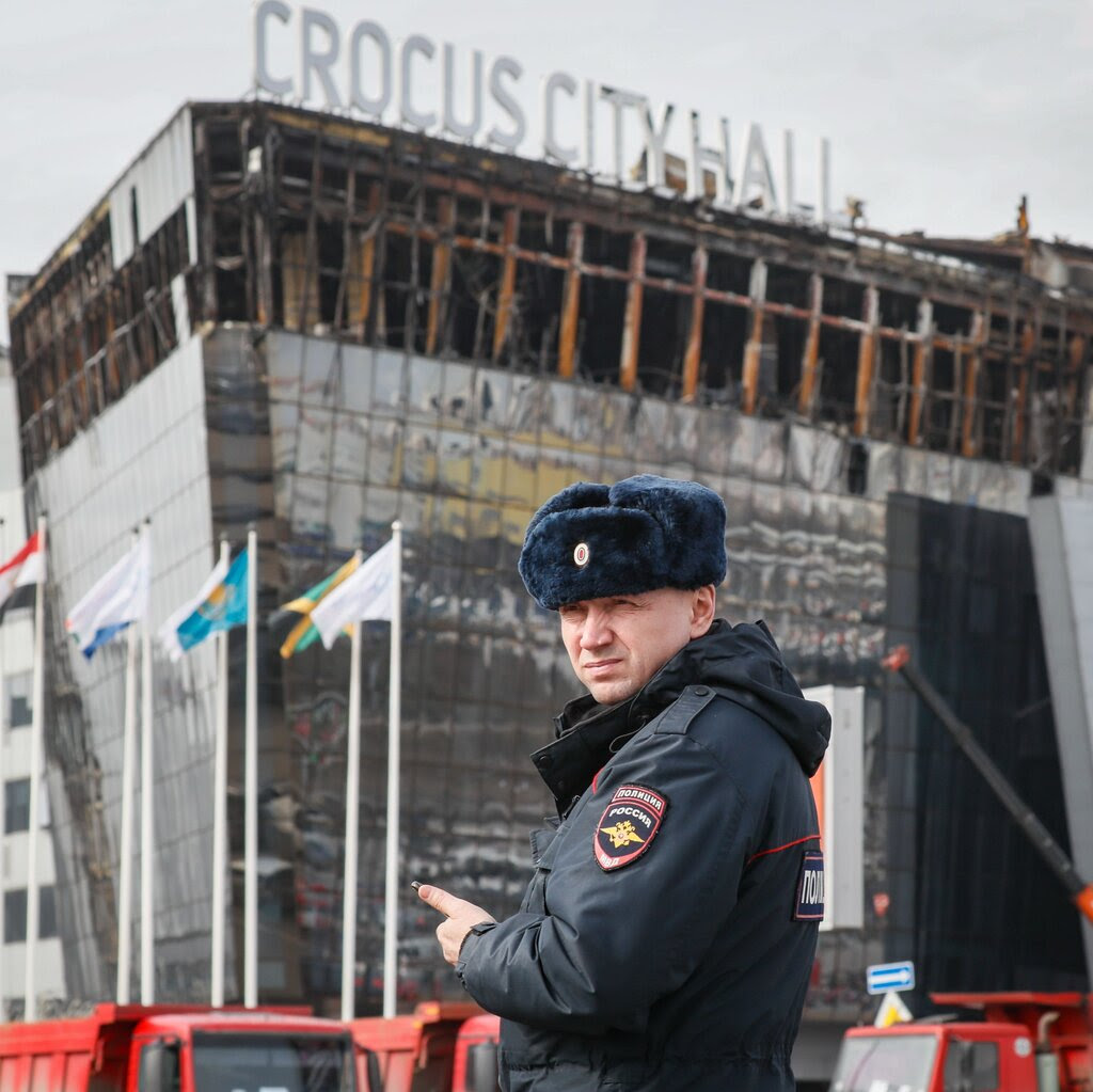 A Russian police officer wearing a fur hat and a dark uniform outside the burned-out Crocus City Hall concert venue.