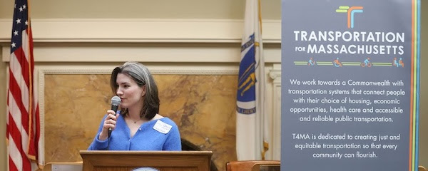Catherine Gleason, a white woman with dark
brown hair and grey and blonde highlights, stands behind a podium. She
is holding a microphone and looking off to the picture's left. Next to
her is a vertical sign with the mission and logo of Transportation for
Massachusetts