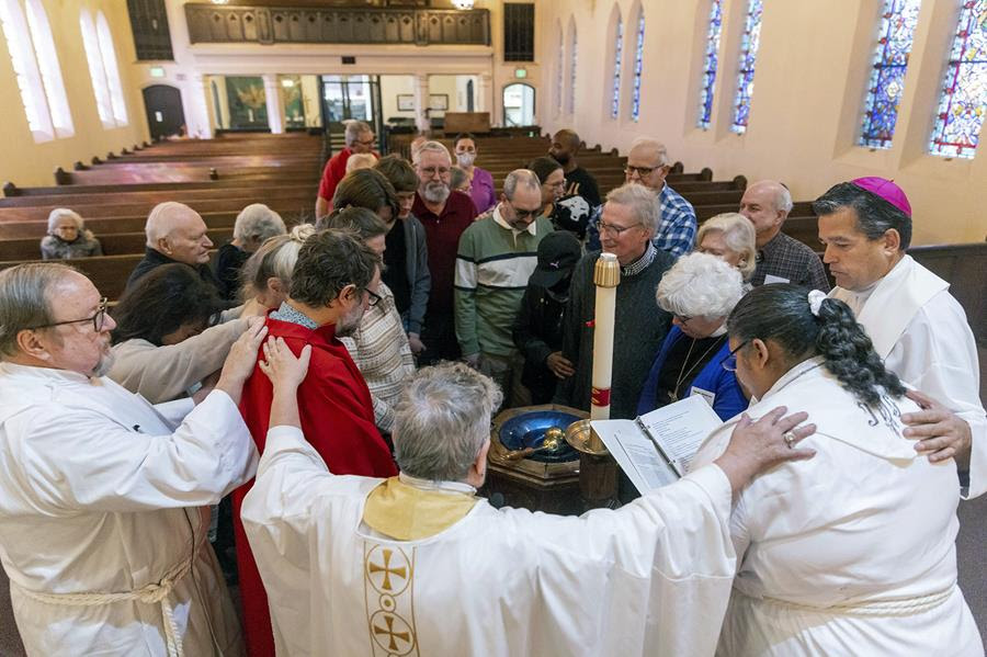 A group of people gather in a circle with their heads bowed in prayer. Some of the people in the circle are wearing religious robes and have their hands laid on the shoulders of the people beside them.