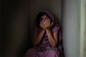 A teenaged girl is sitting in the corner of a dark room. She is covering most of her face with her hands.