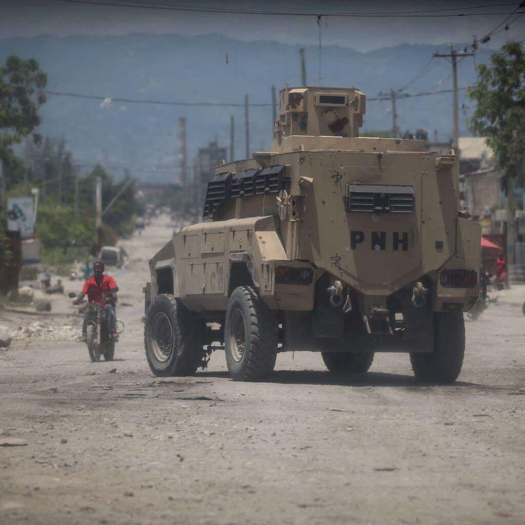 An armored vehicle on a dusty street, where several people can be seen riding motorbikes.