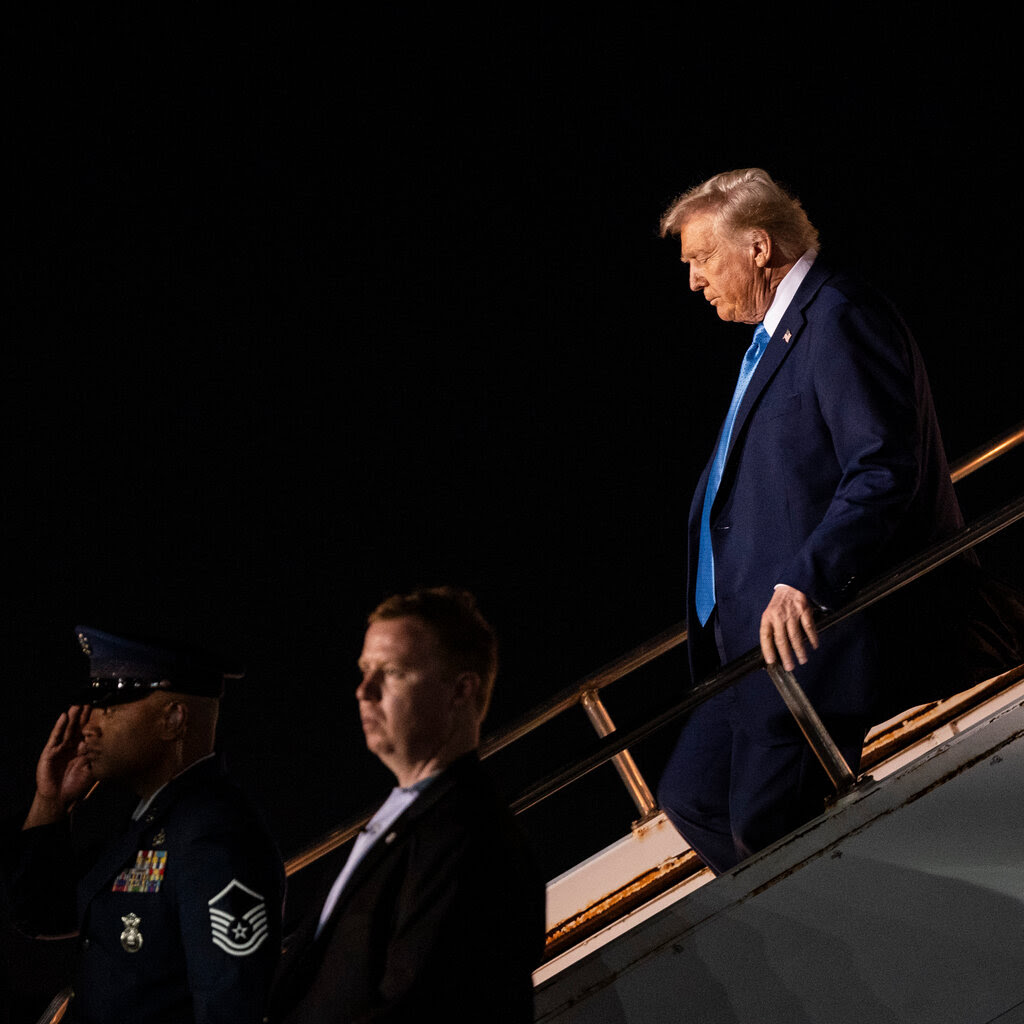 President Trump, walks down the stairs while leaving a plane. Two man, one of them in uniform, stand to his side.