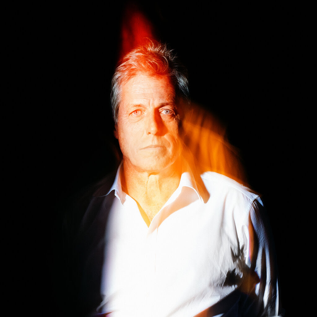 A portrait of the actor Hugh Grant, who is standing in front of a black background with a orange streak of light in front of him.