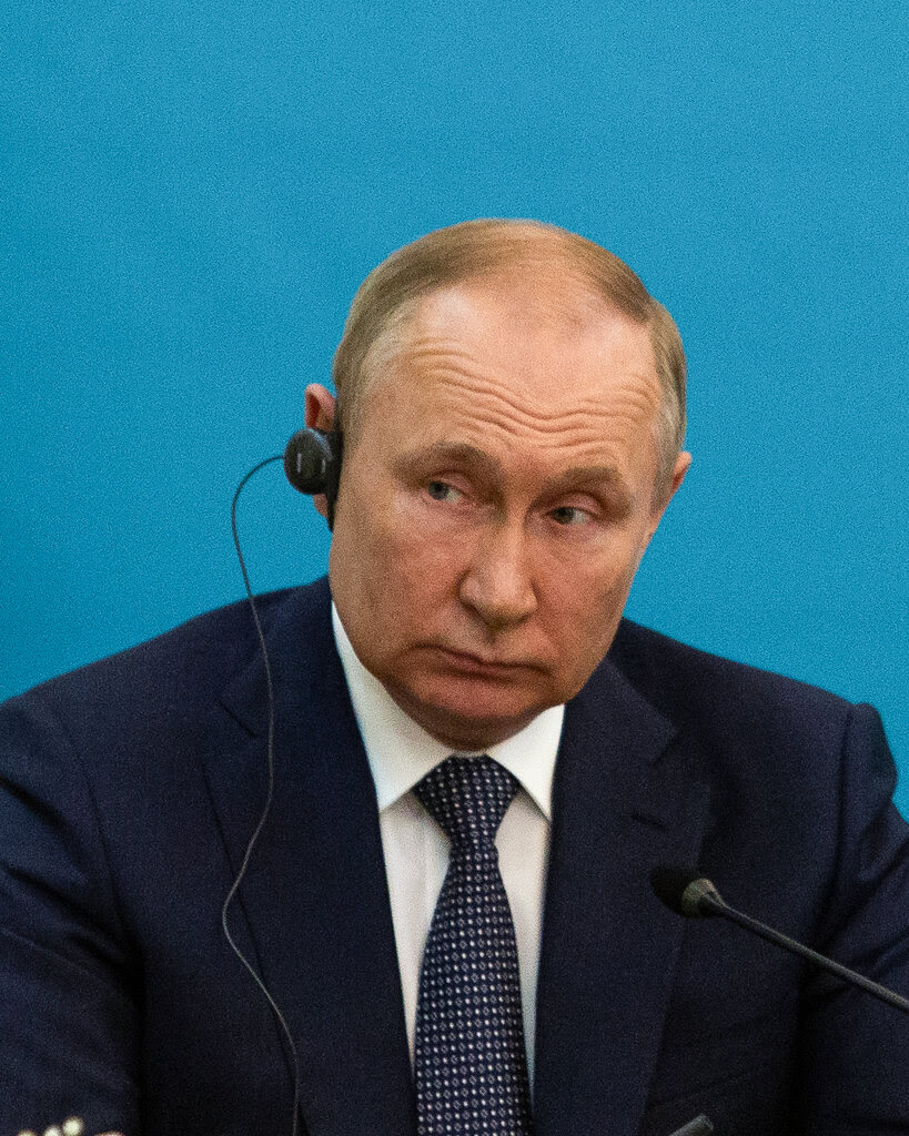 Vladimir Putin in a black suit with a circle and diamond patterned tie sits in front of a microphone while listening to an ear piece in his right ear.
