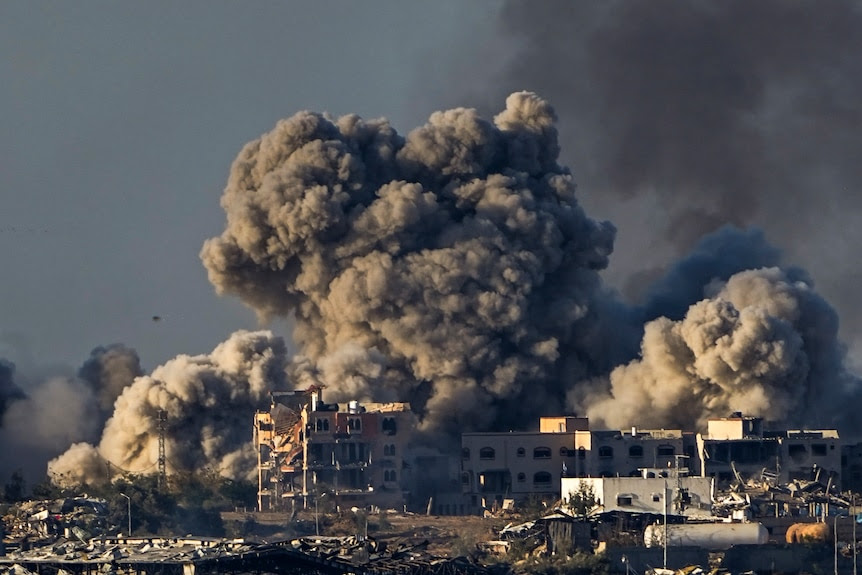 large, thick plume of smoke rises while damaged buildings and rubble can be see in the foreground